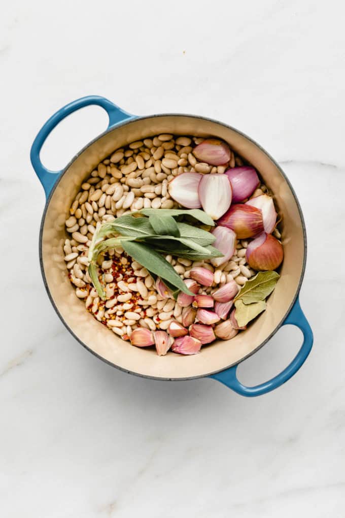 A blue pot filled with white beans, sage, shallots, garlic and bay leaves