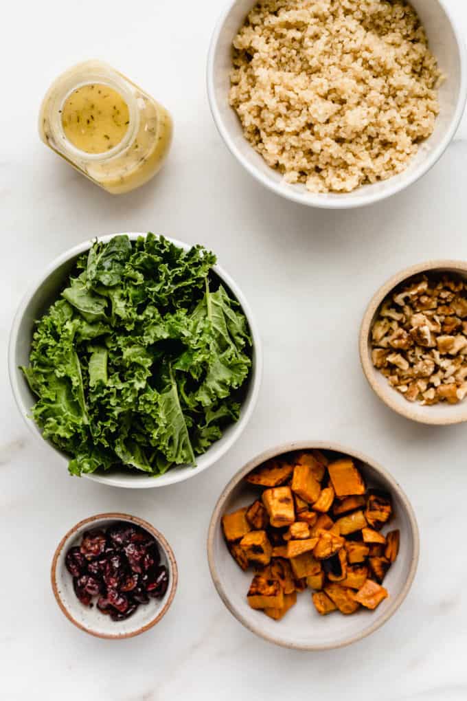 Kale, quinoa, sweet potato, walnuts, cranberries and salad dressing in bowls on a marble baord