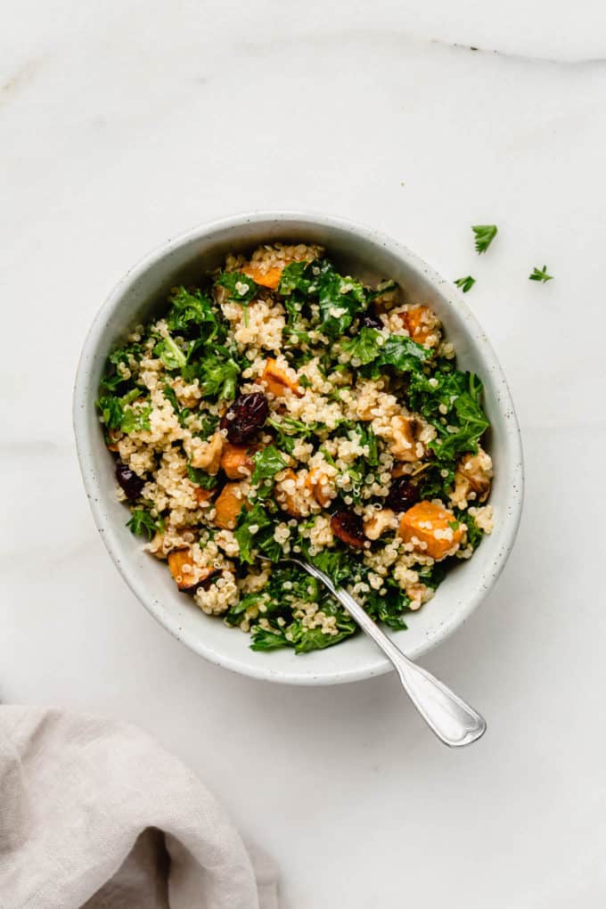 A bowl of quinoa kale salad with sweet potatoes in a white bowl with a napkin on the side
