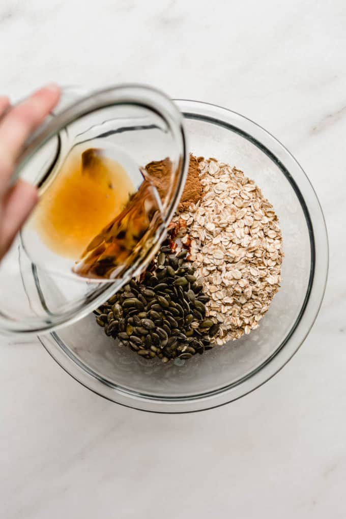 a hand pouring maple syrup into a bowl of oats, pumpkin seeds and pecans