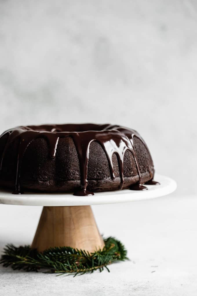 A gingerbread bundt cake topped with chocolate glaze on a marble and wooden cake stand