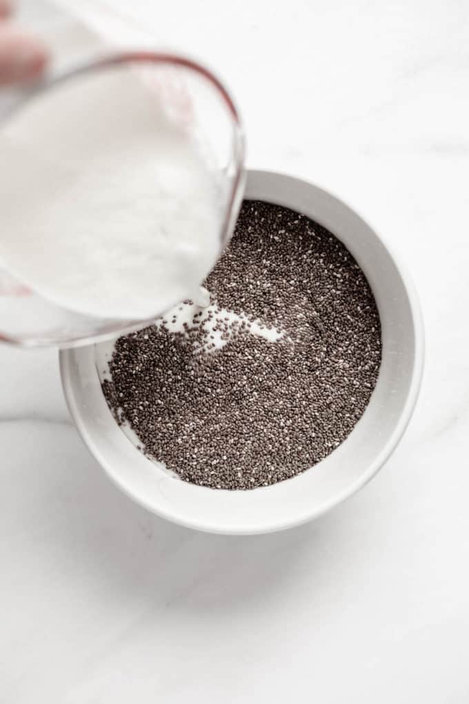 A measuring cup pouring coconut milk into a white bowl with chia seeds in it