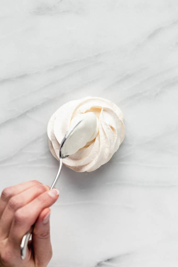 a hand spooning coconut whipped cream into the centre of a pavlova