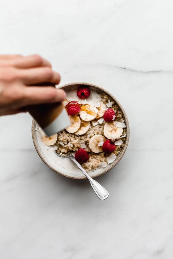 A hand pouring maple syrup onto a bowl of breakfast quinoa