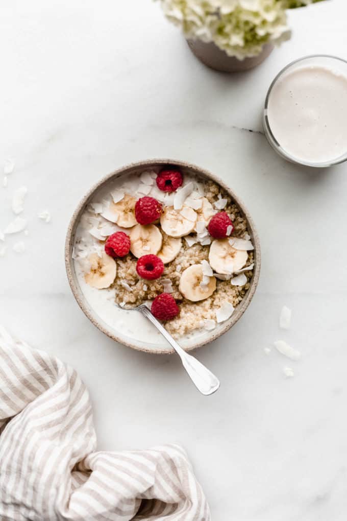 A breakfast quinoa bowl topped with bananas and berries with a glass of milk and flowers on the side