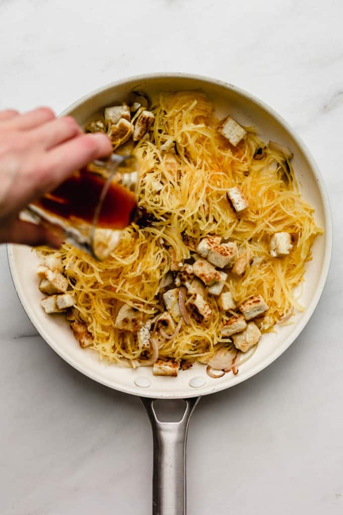 A hand pouring pad Thai sauce into a pan of spaghetti squash and tofu