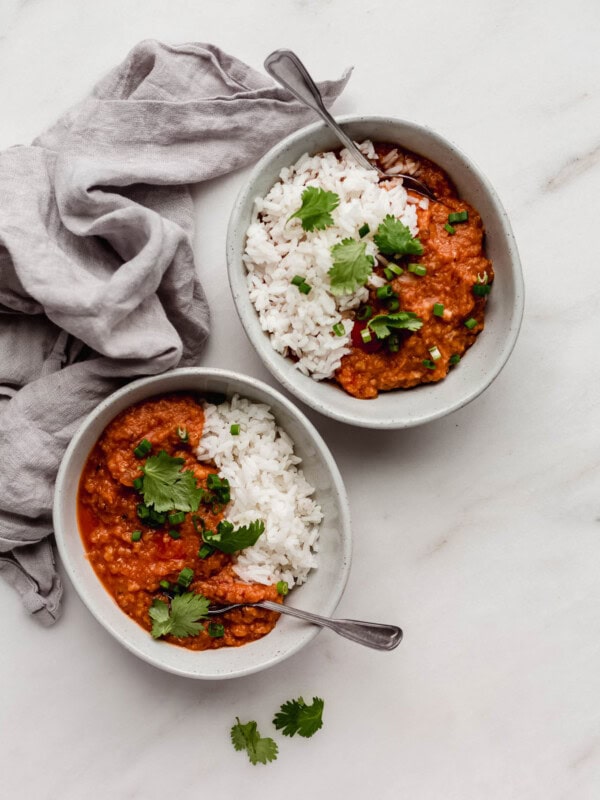 two bowls of red lentil curry and rice with spoons in them and a grey napkin on the side