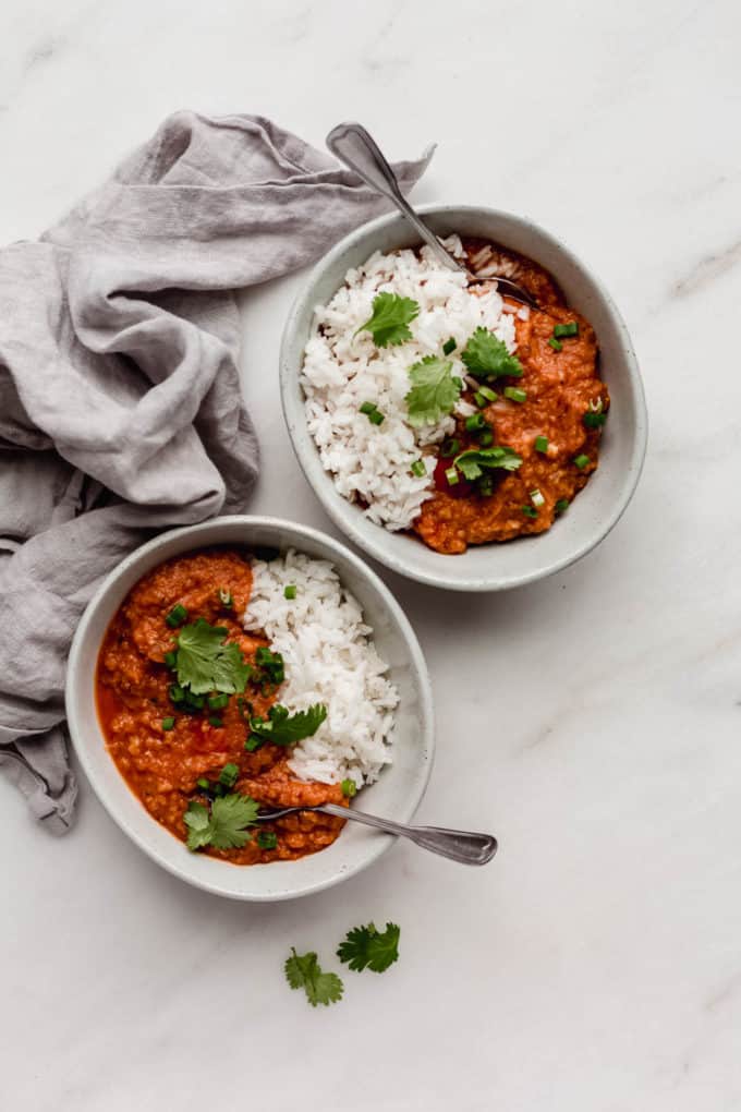 two bowls of red lentil curry and rice with spoons in them and a grey napkin on the side