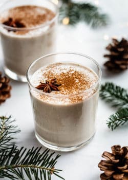 A glass of vegan eggnog on a white counter surrounded by pinecones and pine leaves