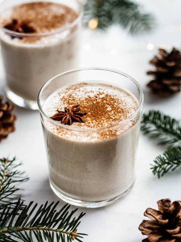 A glass of vegan eggnog on a white counter surrounded by pinecones and pine leaves