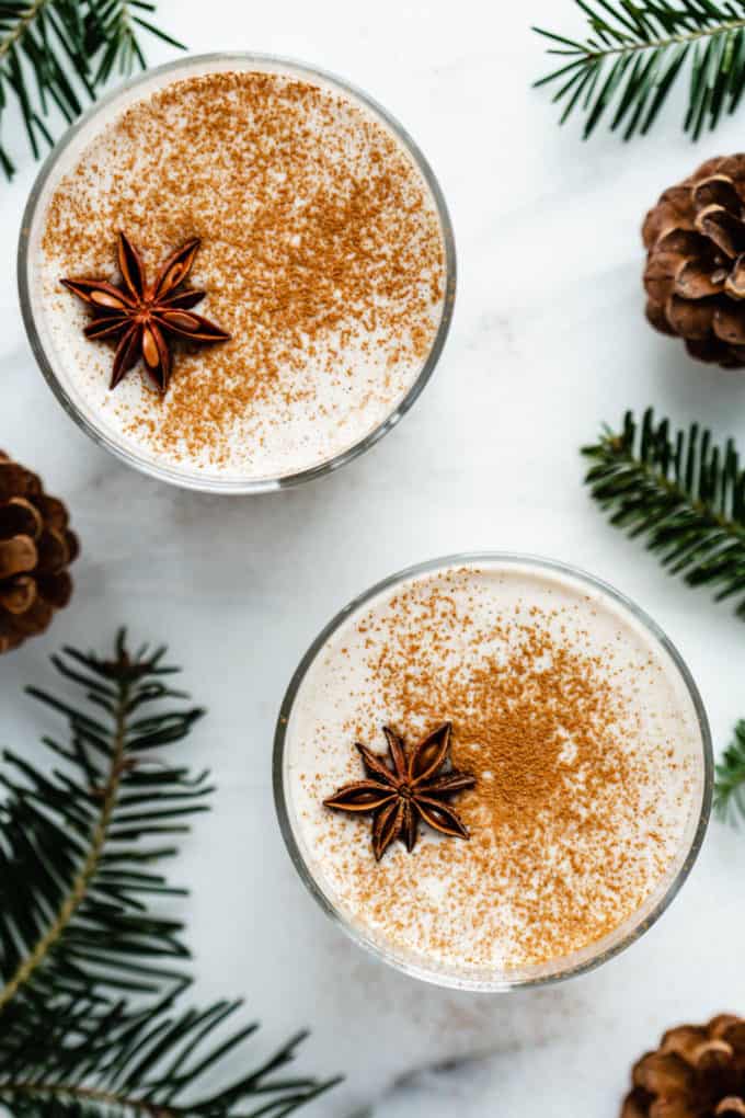 A topdown view of two glasses of vegan eggnog on a white counter with pine leaves around them
