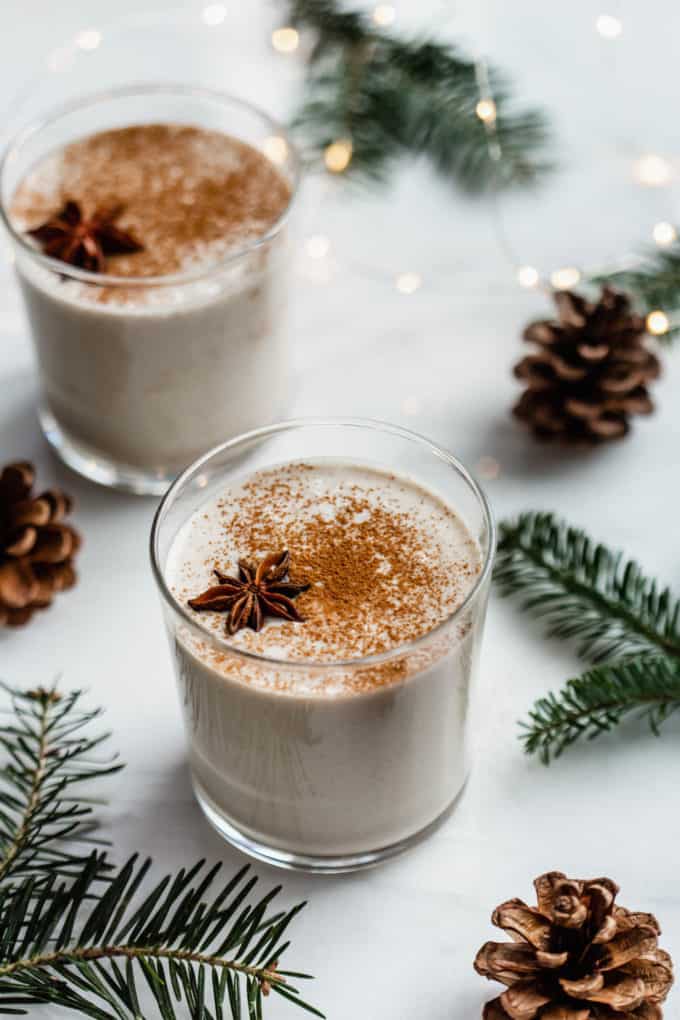 two cups of homemade eggnog topped with grated nutmeg and Christmas lights in the background