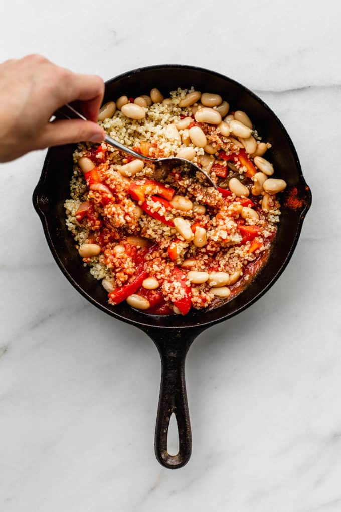 A hand holding a spoon mixing quinoa and beans in a cast iron skillet