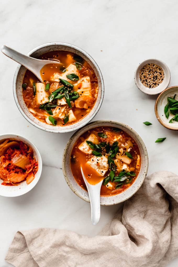 two bowl of kimchi soup with a side of kimchi, sesame seeds and green onions