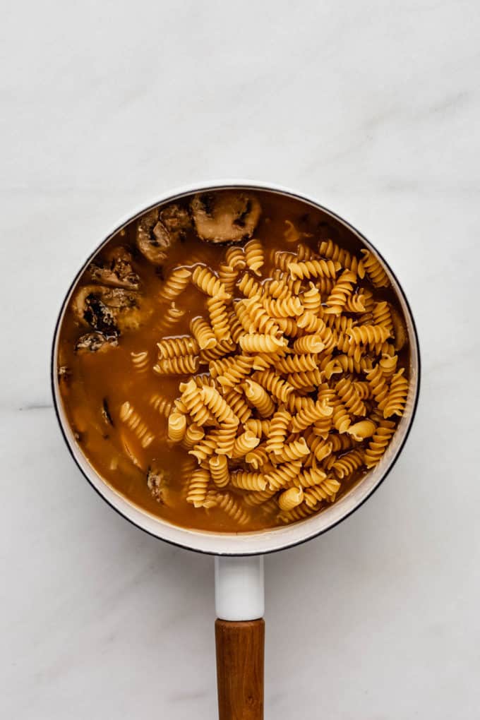 A white pot with mushrooms, broth and uncooked pasta in it