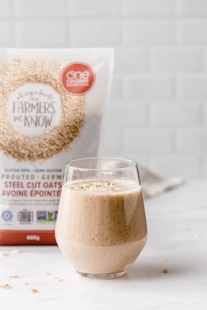 A glass of banana bread smoothie with a pack of steel cut oats in the back