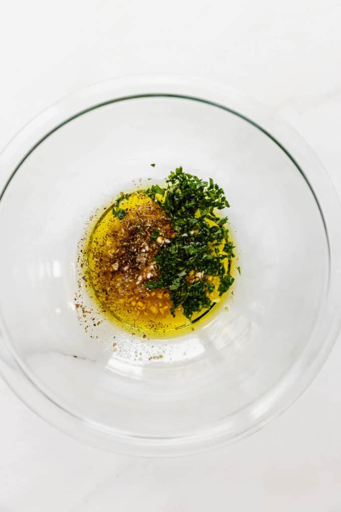 A clear mixing bowl with olive oil, herbs and garlic in it