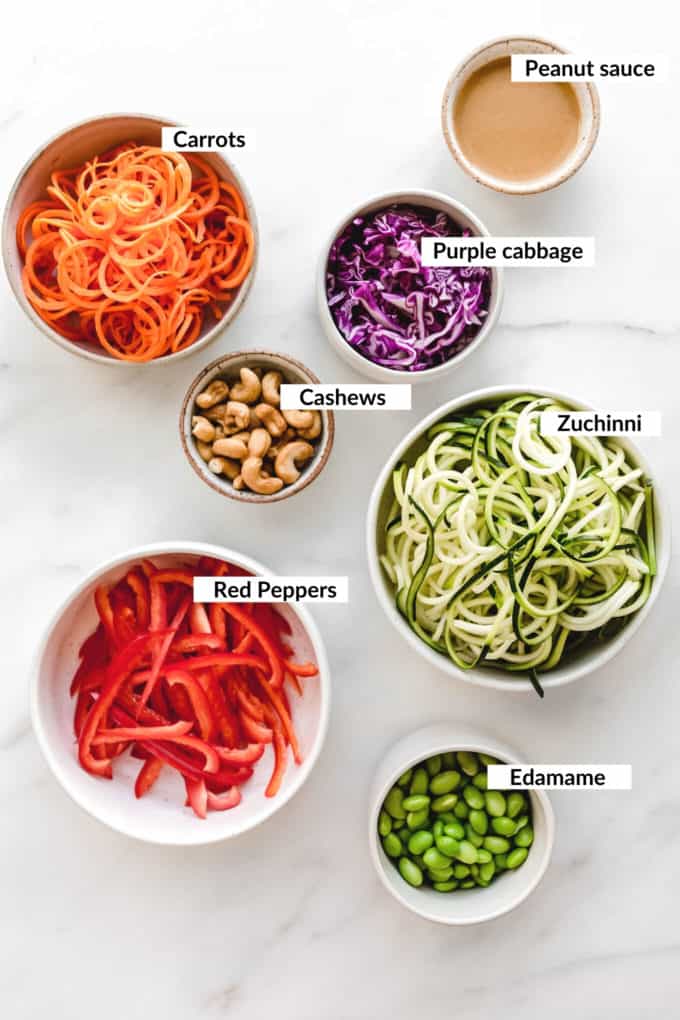 A flat lay of carrots, zucchini, peppers, cabbage, edamame, cashews and peanut sauce in bowls