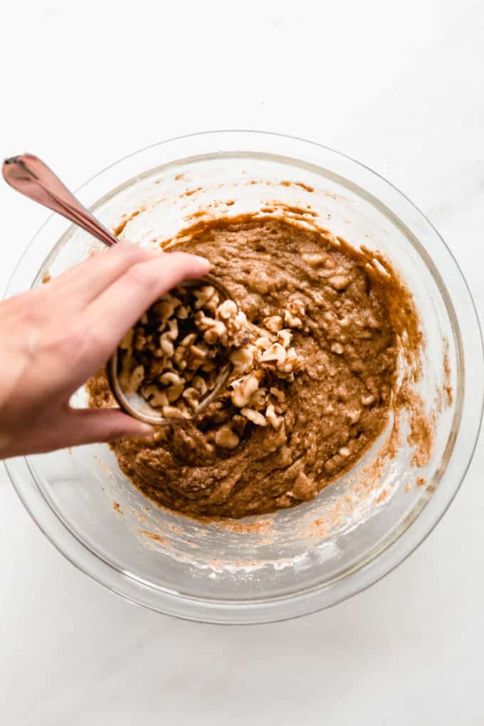 A hand pouring walnuts into a bowl of muffin batter