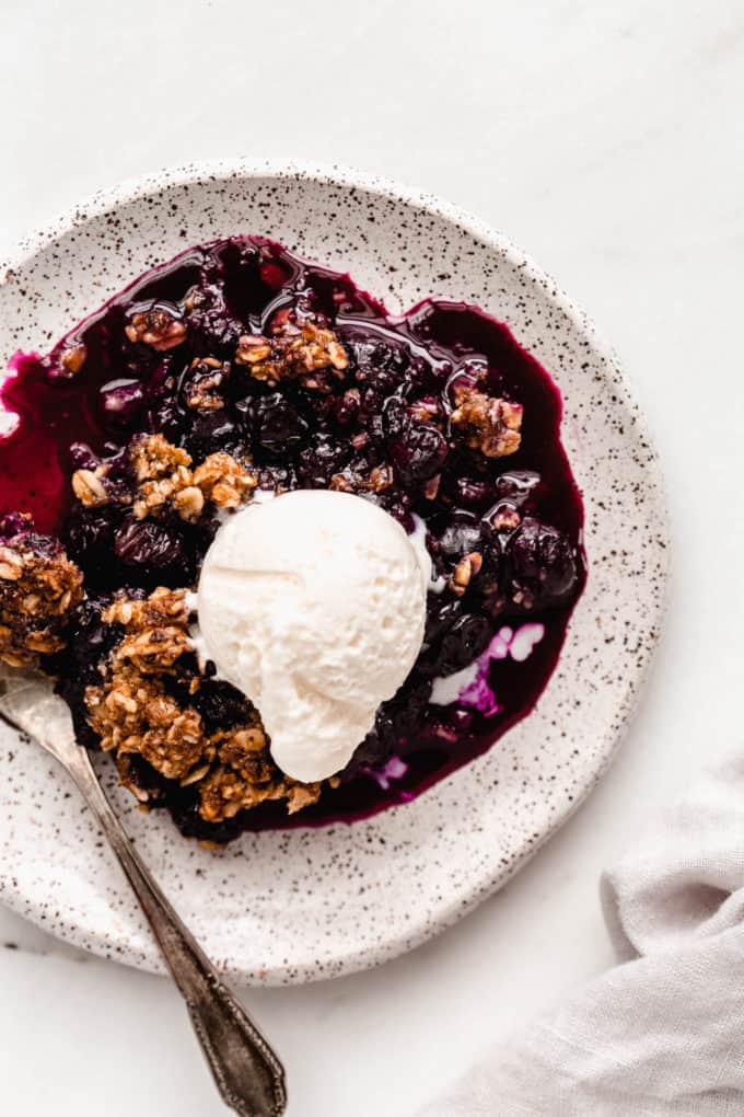 Blueberry crisp on a white speckled plate topped with ice cream