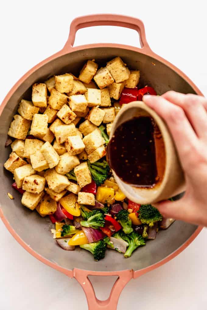 a hand pouring stir fry sauce into a pan of tofu and vegetables