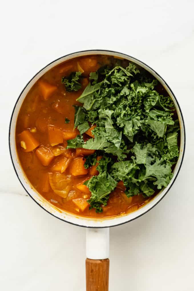 A white pot with squash curry and fresh kale leaves in it