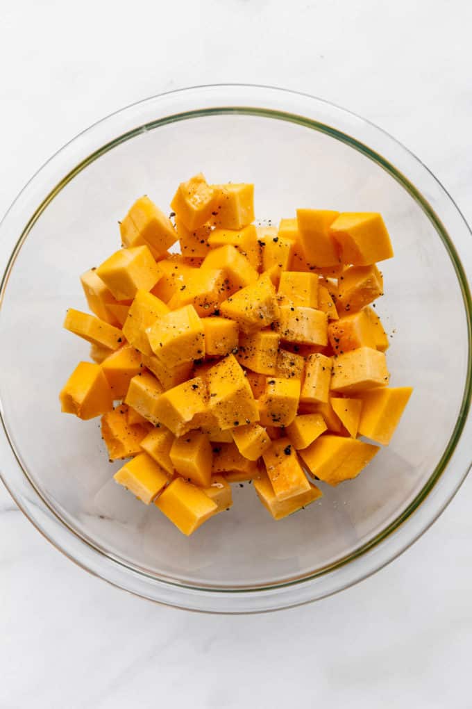 A clear mixing bowl with cubes of pumpkin it it tossed in olive oil, salt and pepper