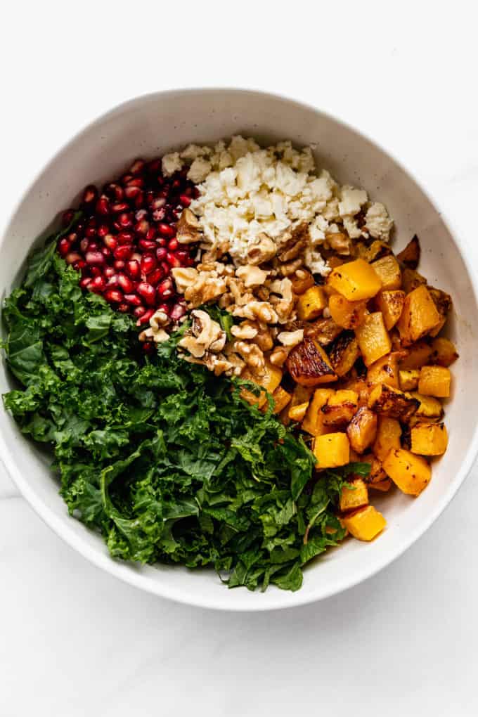 A white mixing bowl with kale, roasted pumpkin, walnuts, feta and pomegranate in it