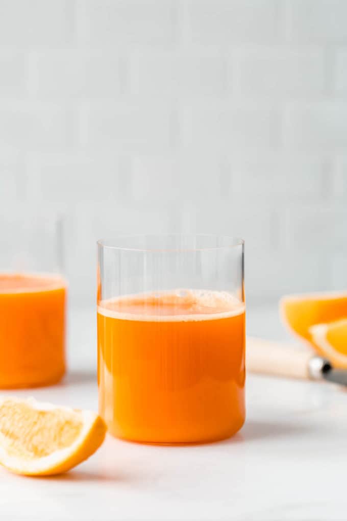 Two glasses or orange and carrot juice on a counter with sliced oranges around them