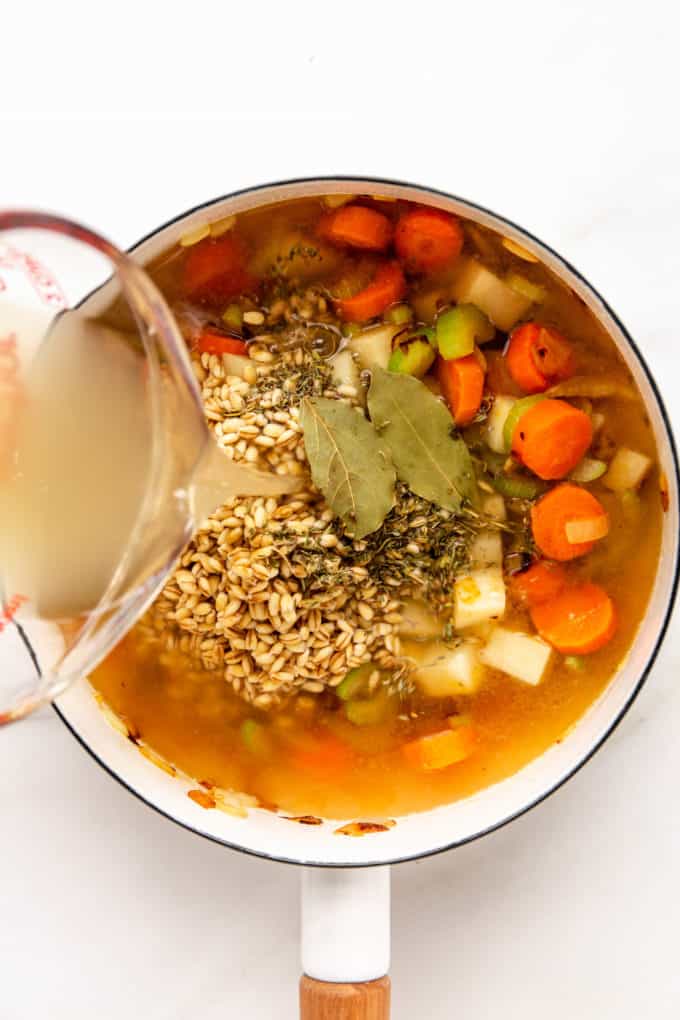 a measuring cup pouring broth into a pot of barley and vegetables