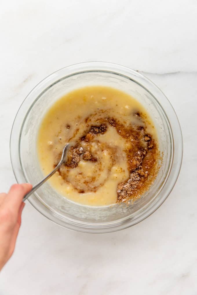 A hand mixing brown sugar into a bowl of mashed bananas