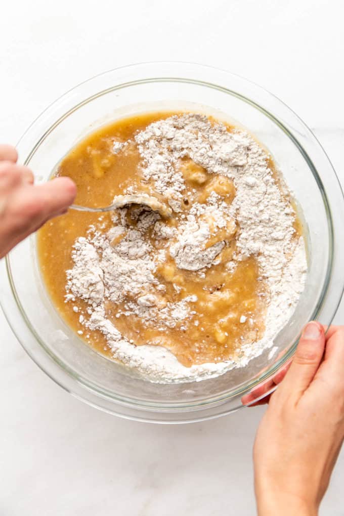 A hand mixing flour with bananas in a mixing bowl