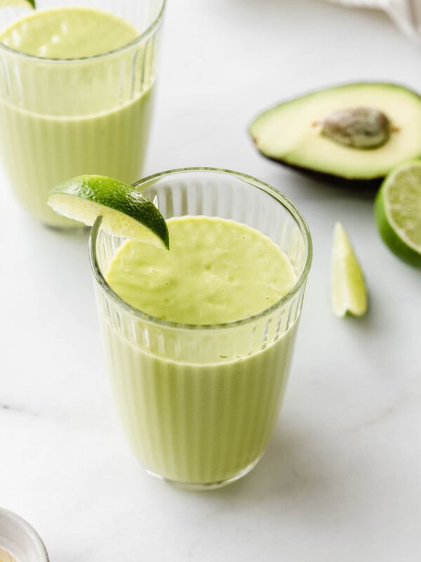 two avocado mango smoothies on a marble counter with sliced avocado and limes