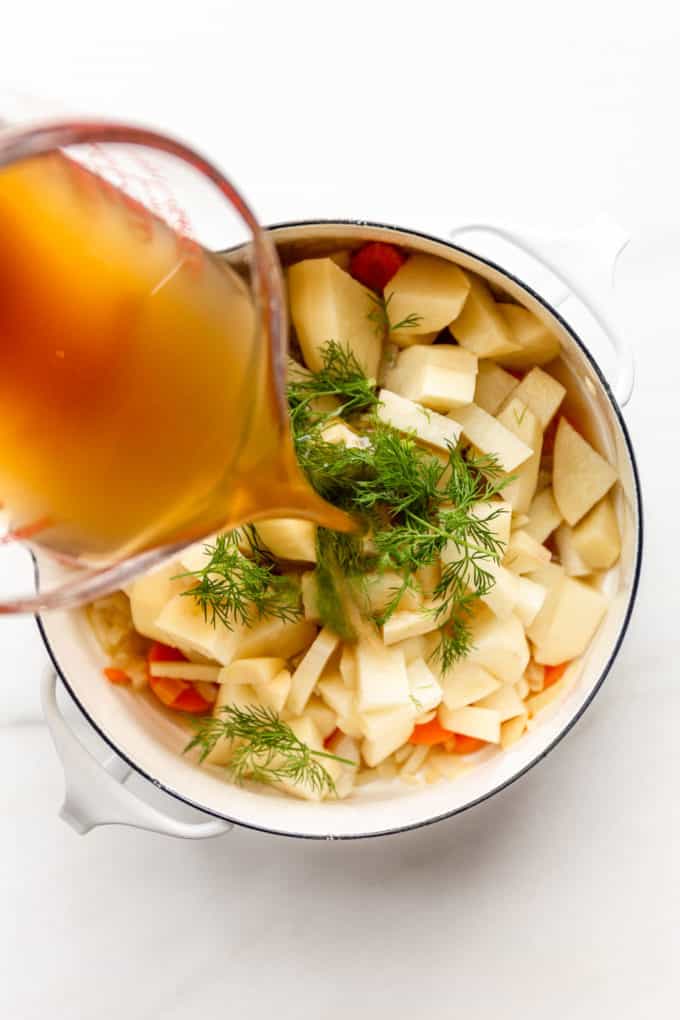 a measuring cup pouring broth into a pot of potatoes and dill