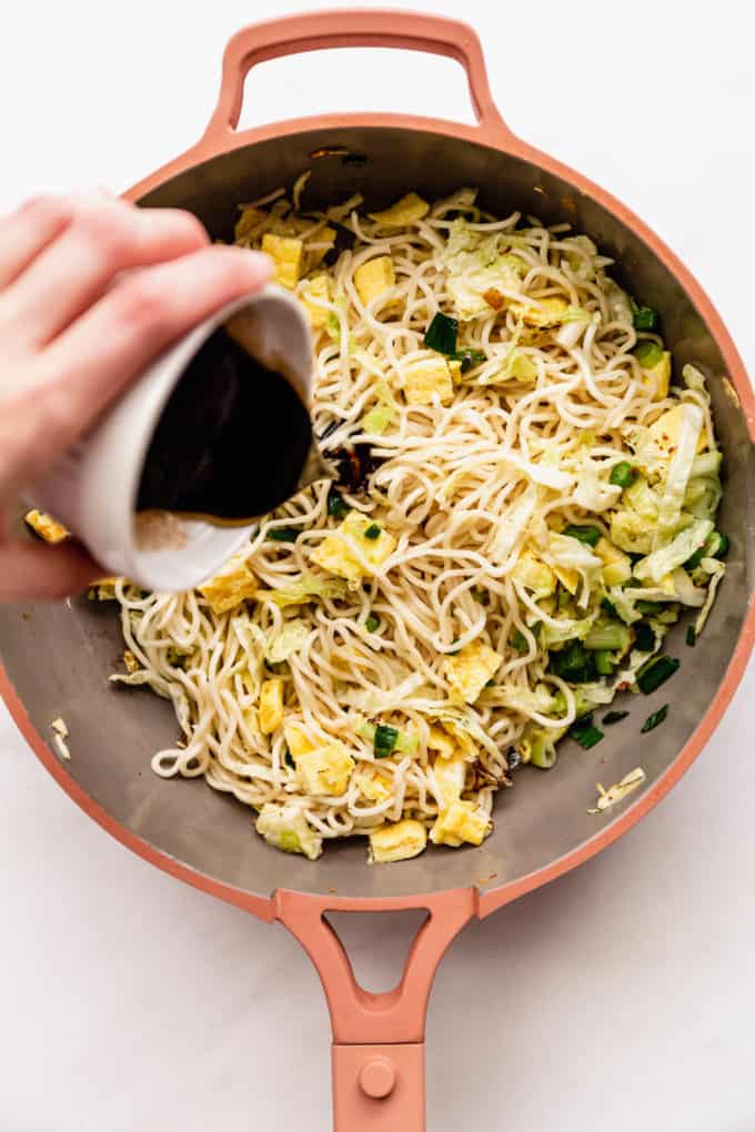 stir-fry sauce being poured into a pan of noodles and vegetables