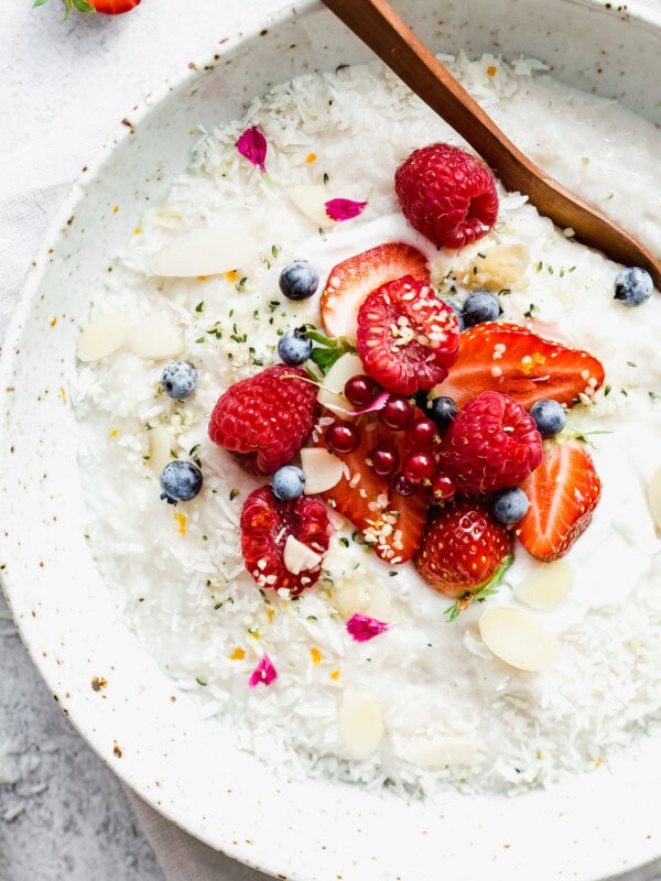 Coconut rice pudding topped with berries, help seeds and shredded coconut in a white speckled bowl