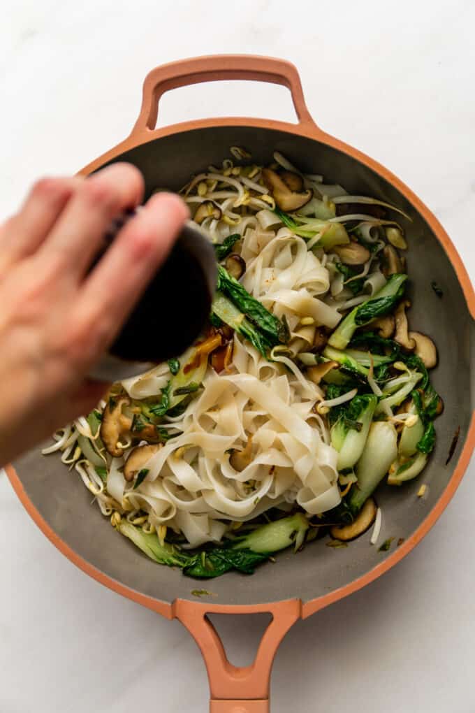 stir-fry sauce being poured into a pan of veggies and rice noodles