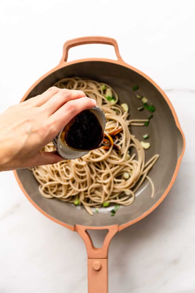 a hand pouring sauce into a pan of noodles