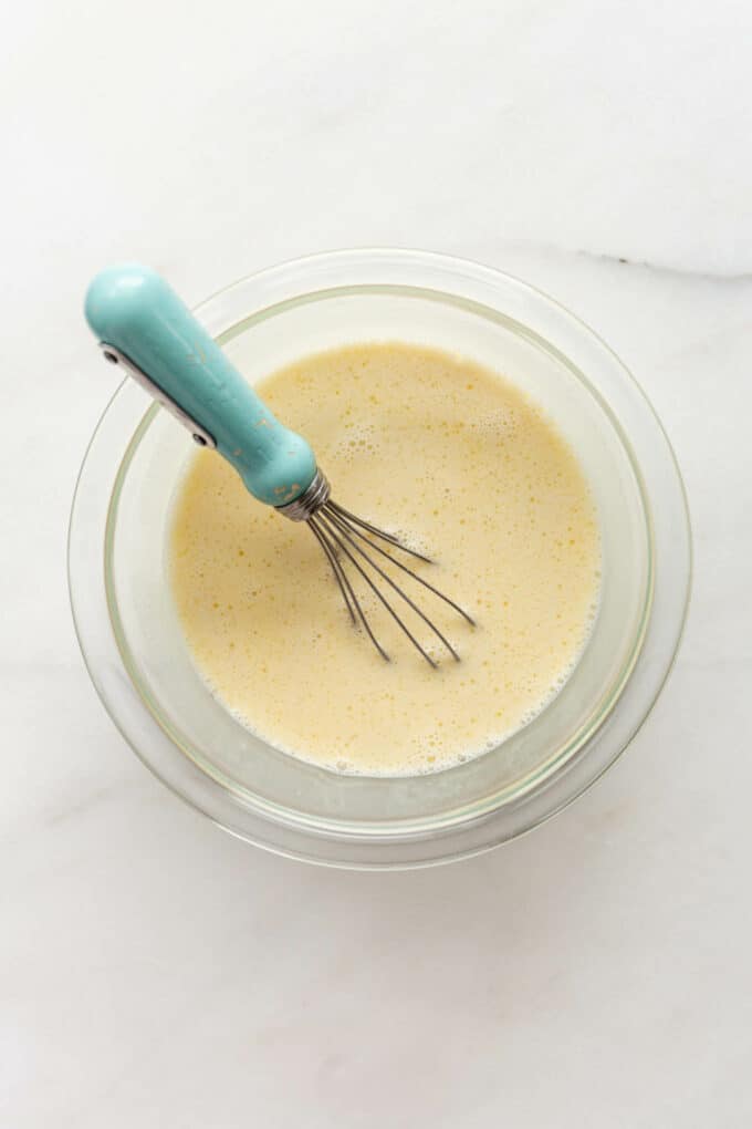 whisked eggs and milk in a clear bowl with a blue whisk