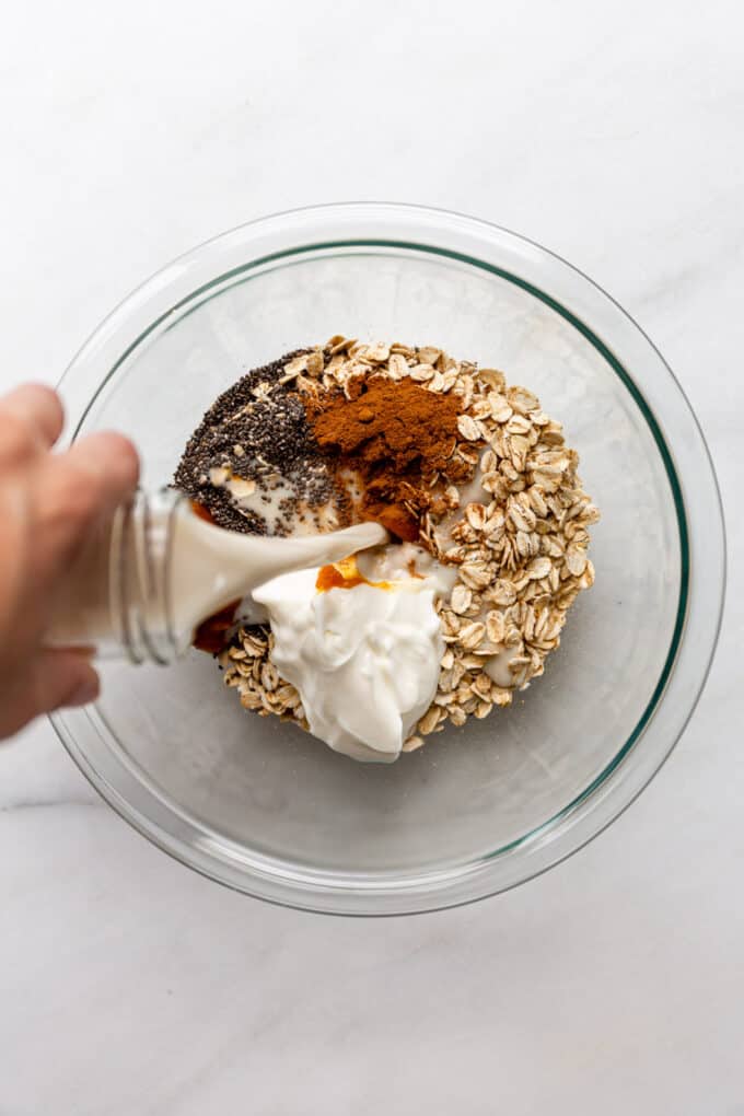 almond milk being poured into a bowl of oats with chia seeds, pumpkin puree, cinnamon and yogurt