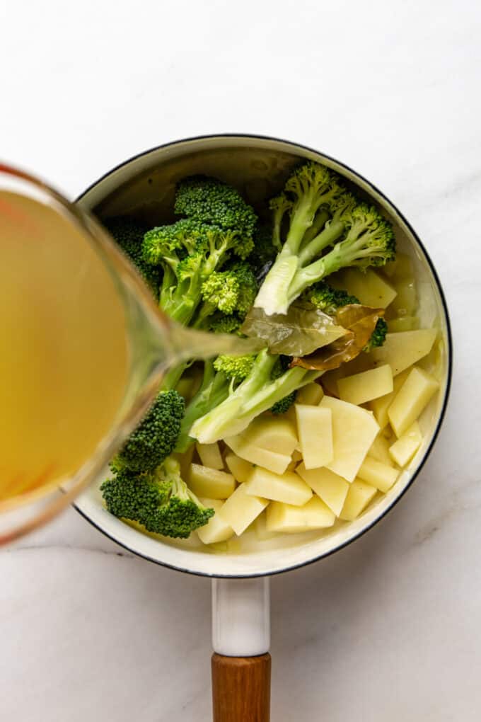a measuring cup pouring broth into a pot of broccoli and potato