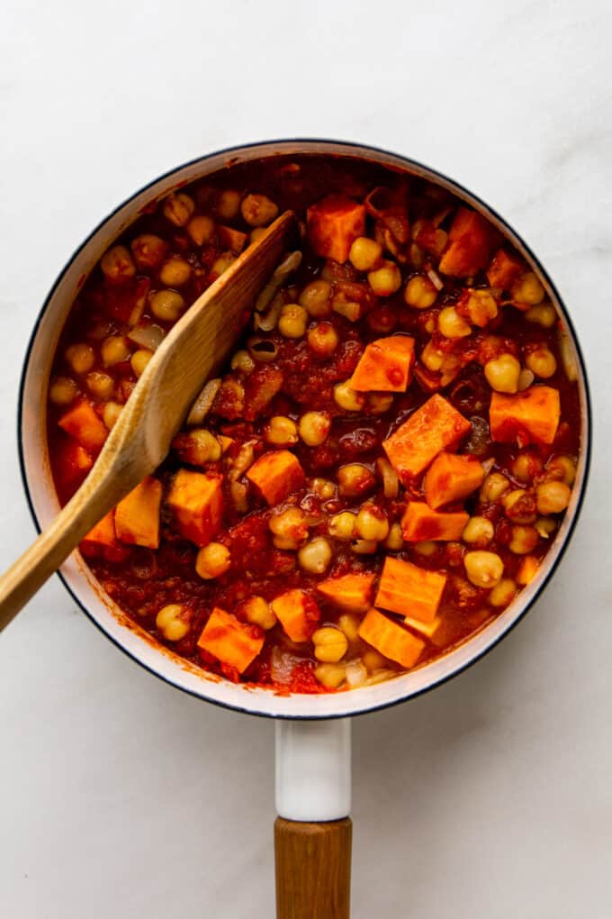 a wood spoon mixing potatoes, chickpeas and tomato puree in a white pot