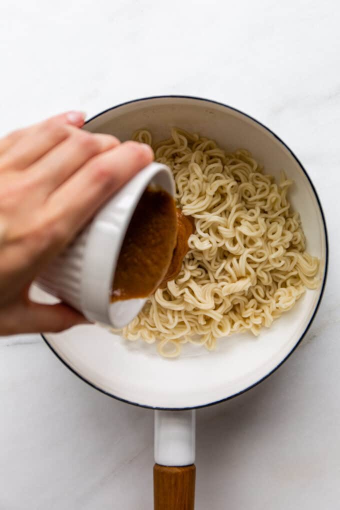 a hand pouring peanut butter sauce into a pot of ramen noodles