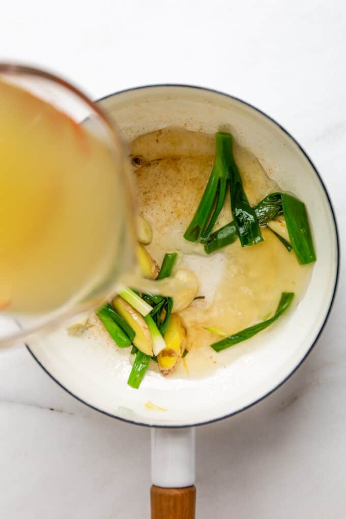 vegetable broth being poured into a white pot with scallions, ginger and garlic
