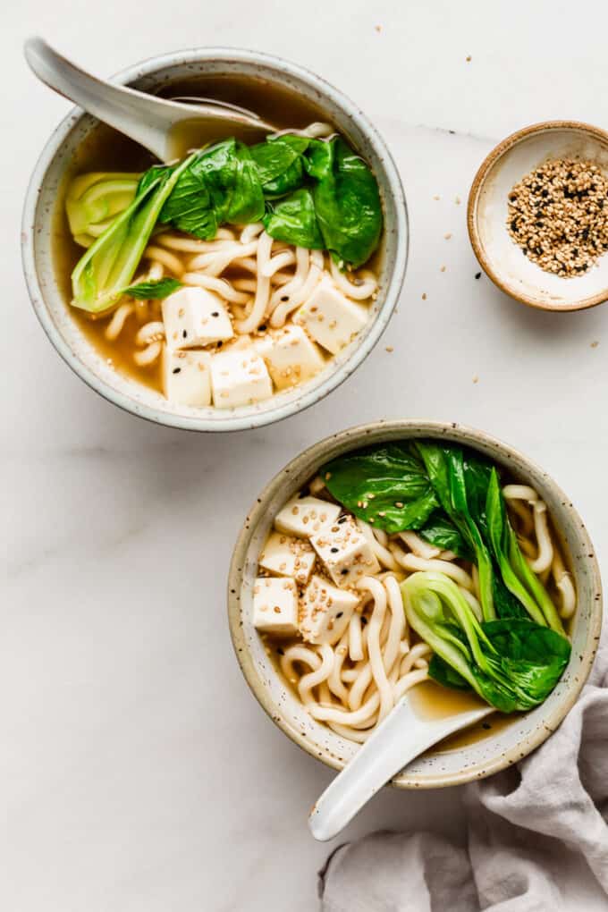 two bowls of udon noodle soup with a small bowl of sesame seeds on the side