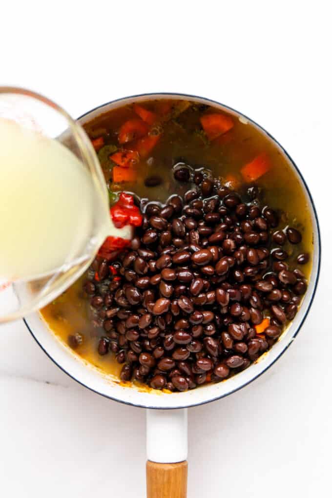 broth being poured into a pot of black beans and vegetables