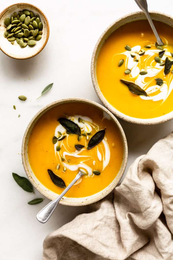 two bowls of acorn squash sup with a napkin and small bowl of pumpkin seeds on the side