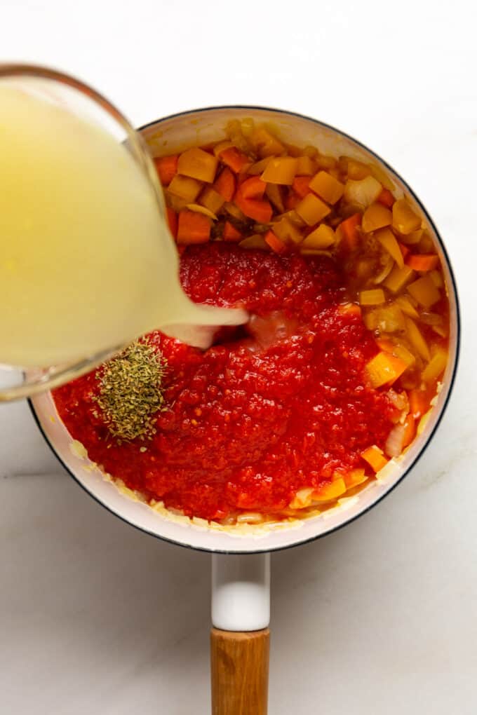 broth being poured into a pot of crushed tomatoes and chopped vegetables
