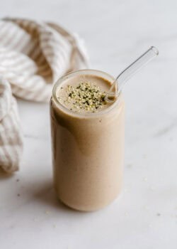 A banana date smoothie in a glass topped with hemp seeds and a striped beige napkin on the side.