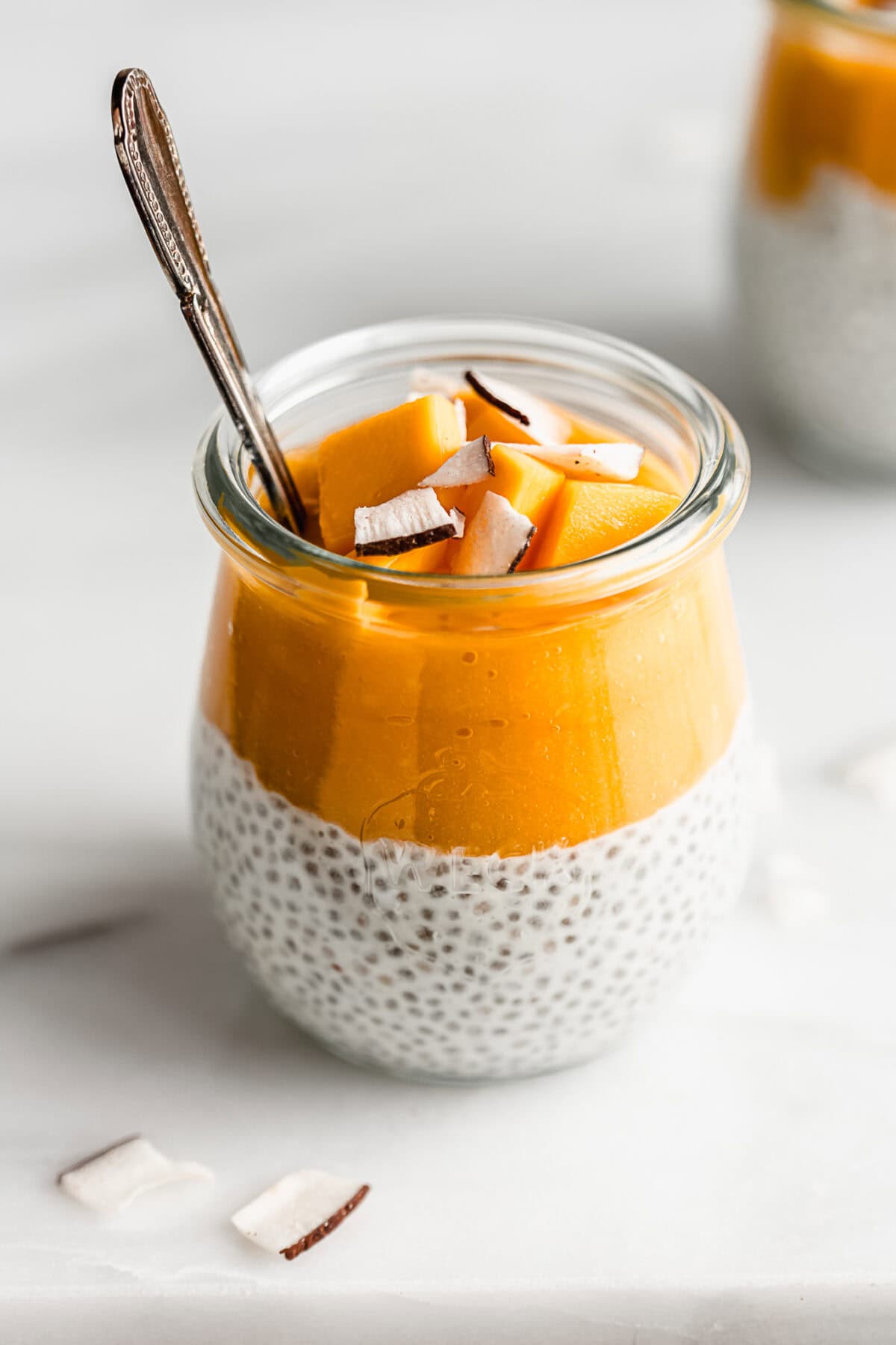 A cup of coconut chia pudding topped with mango puree, chunks of mango and coconut flakes.